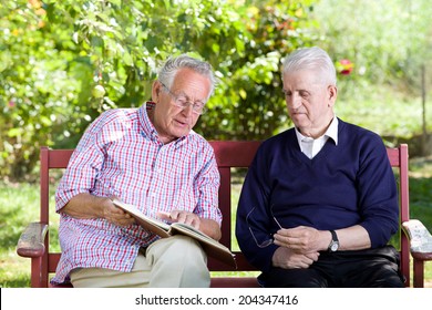 Two Senior Men Sitting In Park And Reading A Book