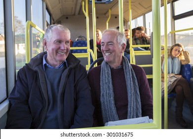 Two Senior Men Smiling And Talking On The Bus. There Are Other People Sitting On The Bus In The Background. 