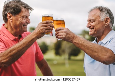 Two Senior Men Toasting Beer Glasses Outdoors. Smiling Mature Male Friends Cheering Beers While Standing Outside.