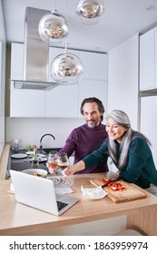 Two Senior People Making Video Call On Laptop In Modern Kitchen While Drinking Wine. Talking And Smiling, Breakfast Time Concept. Stock Photo