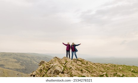 Two senior people stand on a rocky hilltop with arms outstretched, enjoying the view. Overcast sky, rocky terrain, and nature landscape. Two seniors enjoying the mountain view. Seniors travelling. - Powered by Shutterstock