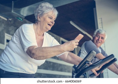 Two Senior People Working Out On Elliptical Machine. Seniors Workout In Gym. Senior Woman Using Smart Phone.