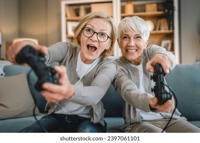 Two senior women caucasian friends or sisters happy old siblings pensioner playing video game console using joystick or controllers while sitting at home real people family leisure concept copy space - Powered by Shutterstock
