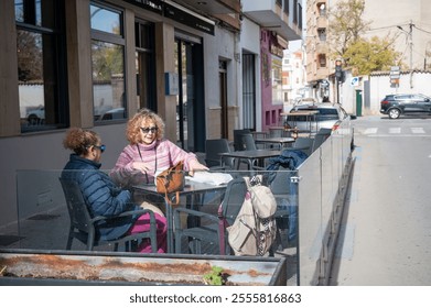 Two senior women enjoying a casual conversation at an outdoor café table in a rural Spanish town - Powered by Shutterstock