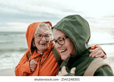 Two senior women laughing together on windy beach in raincoats - Powered by Shutterstock