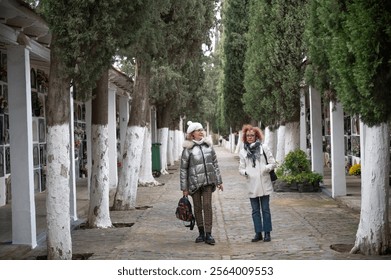 Two senior women walking and talking in a tree-lined cemetery pathway during a calm autumn day, reflecting on memories surrounded by graves and greenery - Powered by Shutterstock