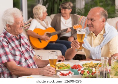 Two Seniors Drinking Beer, In The Background Elderly Women Playing Guitar