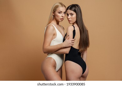 Two sensual elegant women in swimsuits posing together isolated over beige background
