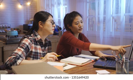 two serious asian woman discussing on project report for final exam. girl pointing laptop screen showing classmate something talking. group of college students teamwork on kitchen table in dark home. - Shutterstock ID 1351649420