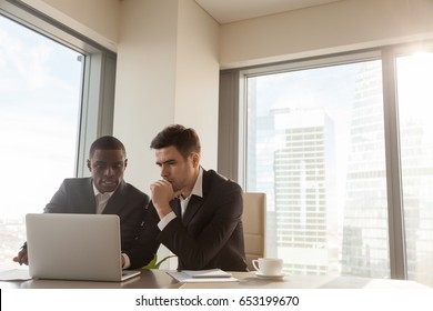 Two serious black and white businessmen discussing project sitting at office desk, using pc laptop, looking at screen, make presentation, create business plan, fulfill task, consider investment 
