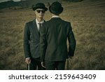 Two serious calm men in black suits, bowler hats and black sunglasses stand facing each other in an open wild field. Surrealist style.