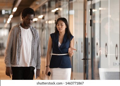 Two serious diverse colleagues talking walking in modern office hallway, asian female and african black male professionals discussing project having business conversation going in work space hall