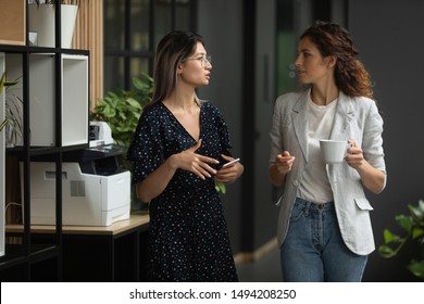 Two serious diverse female colleagues team talk walk in modern office, asian businesswoman having business conversation with caucasian coworker discuss project work meeting in company hallway space