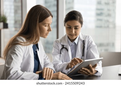 Two serious multiethnic doctor colleagues working with tablet together, reviewing electronic medical records, talking, looking at screen, using medical application, modern technology for job - Powered by Shutterstock