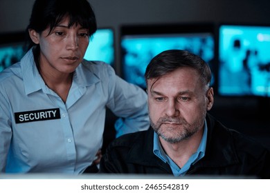 Two serious security guards looking at computer screen in control room while observing situation in supermarket or some other building