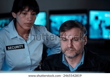 Two serious security guards looking at computer screen in control room while observing situation in supermarket or some other building