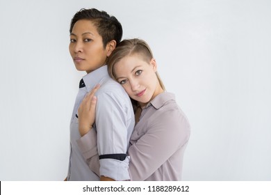 Two Serious Women Hugging And Looking At Camera. Multiethnic Homosexual Couple Posing. Lesbian Couple Concept. Isolated Side View On White Background.