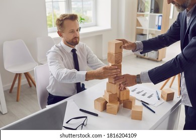 Two Serious Young And Old Businessmen Building Wood Block Tower In Company Office As Metaphor For Business Succession And Collaboration. Concept Of Risk Management And Maintaining Financial Stability