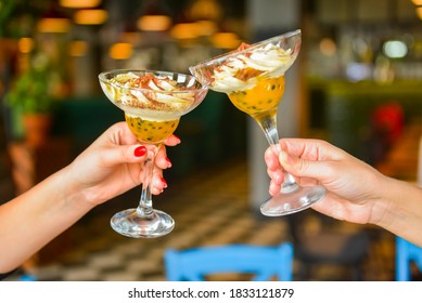 Two Servings Of Chocolate Cream Dessert In Tall Martini Glasses. Two Young Women Holding Glasses With Dessert Over Blurred Restaurant Background, Selective Focus. Cheers, Leisure Time With Friends.