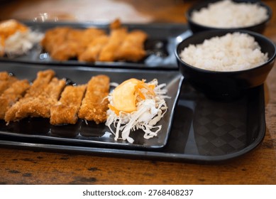 Two sets of sliced breaded meat with shredded cabbage and two bowls of white rice are arranged on a black patterned tray. The tray sits on a wooden textured table.