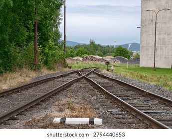 Two sets of train tracks split and extend toward distant hills, bordered by green trees and gravel piles. A concrete wall and utility poles line the right side