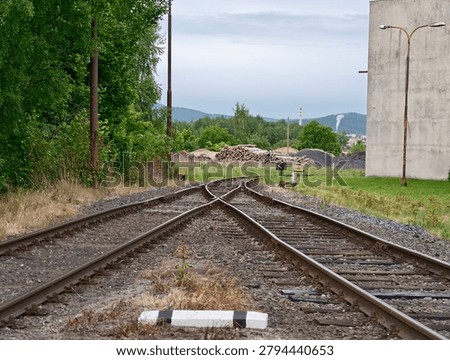Two sets of train tracks split and extend toward distant hills, bordered by green trees and gravel piles. A concrete wall and utility poles line the right side