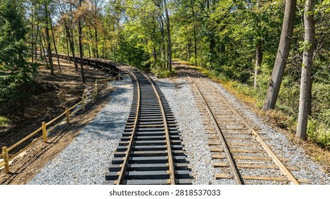 Two sets of train tracks stretch through a wooded area with green and brown leaves. The tracks curve along the path, surrounded by gravel and wooden fencing. Sunlight shines on the scene.