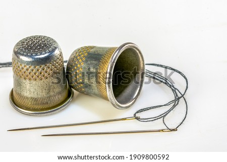 Two sewing needles with black thread and two sewing thimbles lie on a white background