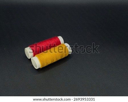 Two sewing thread spools in red and yellow standing side by side on a dark surface, photographed under soft studio light.