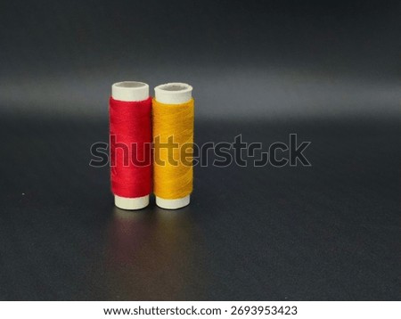 Two sewing thread spools in red and yellow standing side by side on a dark surface, photographed under soft studio light.