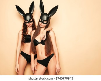 Two sexy beautiful women wearing carnival black mask of Easter bunny rabbit.Hot brunette girls posing near wall in studio. Seductive models in nice lingerie