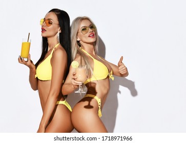 Two Sexy Slim Beautiful Girls In Yellow Bikini Party Hold Cocktails And Juices Smiling Laughing Show Thumb Up On White Background. Summer Vibes Concept
