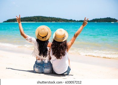 Two Sexy Young Girls, Best Friends Sitting Together On White Sand Beach With The Sun Setting Behind Them With Warm Orange Light, Having Fun .Wearing Straw Hat And Sunglasses. Photographed From Behind