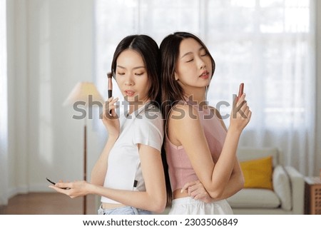 Two shapely young adult Asian models confidently hold lipstick and makeup as they look at the camera with seductive eyes. personal beauty routine. Skin Care, makeup and self-care concept