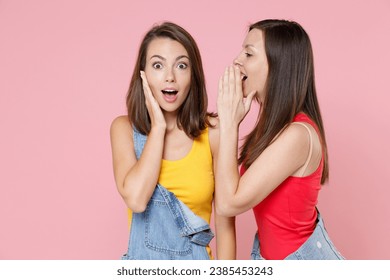 Two shocked amazed young women friends 20s in casual denim clothes posing whispering secret behind hand, sharing news put palm on cheek isolated on pastel pink colour background, studio portrait - Powered by Shutterstock