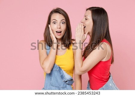 Two shocked amazed young women friends 20s in casual denim clothes posing whispering secret behind hand, sharing news put palm on cheek isolated on pastel pink colour background, studio portrait