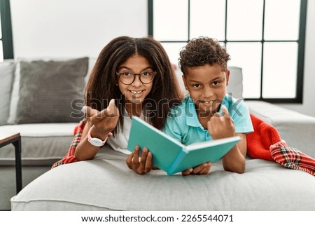 Two siblings lying on the sofa reading a book beckoning come here gesture with hand inviting welcoming happy and smiling 