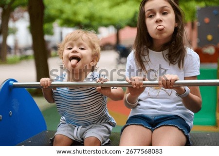 Two siblings pose playfully while sticking out their tongues, seated at a playground under a vibrant green tree. Their joyful expressions reflect carefree childhood moments.