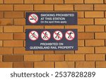 Two signs on a brick wall, one prohibiting smoking and the other forbidding various activities on Queensland Rail property