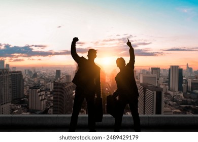 Two silhouetted businessmen celebrate their success on a rooftop with a city skyline and sunset in the background. - Powered by Shutterstock