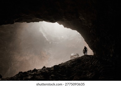 Two silhouetted explorers standing inside massive cave chamber with misty light. The powerful contrast between the pitch-black foreground and the glowing atmospheric haze creates a deeply cinematic and emotionally compelling scene. A striking visual metaphor for human insignificance against nature, discovery, adventure, and the unknown. Ideal for editorial use, travel campaigns, adventure brands, book covers, and inspirational content.