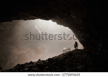 Two silhouetted explorers standing inside massive cave chamber with misty light. The powerful contrast between the pitch-black foreground and the glowing atmospheric haze creates a deeply cinematic and emotionally compelling scene. A striking visual metaphor for human insignificance against nature, discovery, adventure, and the unknown. Ideal for editorial use, travel campaigns, adventure brands, book covers, and inspirational content.