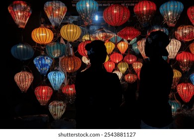 Two silhouetted figures stand in front of a vibrant display of colorful lanterns in a night market in Vietnam, showcasing the illuminated beauty of traditional lanterns. - Powered by Shutterstock