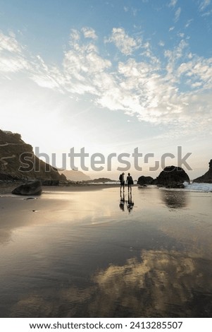 Two silhouetted figures stand near the water edge at Benijo Beach, Tenerife, reflecting on the wet sand during the tranquil sunset