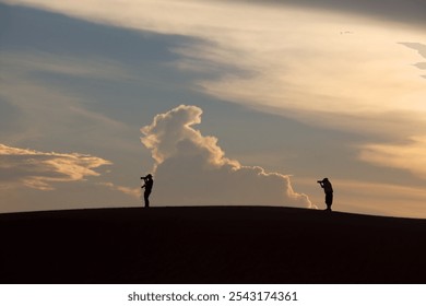 Two silhouetted photographers capturing the stunning sunset over a desert landscape, showcasing vibrant clouds and a serene atmosphere filled with natural beauty. - Powered by Shutterstock