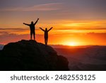 Two silhouettes of happy couple are standing with open arms on big rock against sunset sky and sun