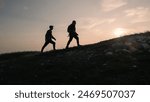 Two silhouettes, male and female, conquering a mountain peak, giving high fives and enjoying the landscape during a romantic sunset, aerial shot.