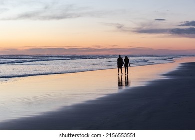 Two silhouettes of unrecognisable people walking along the seashore at sunset. Pink colours of the sky and the reflection in the water. - Powered by Shutterstock