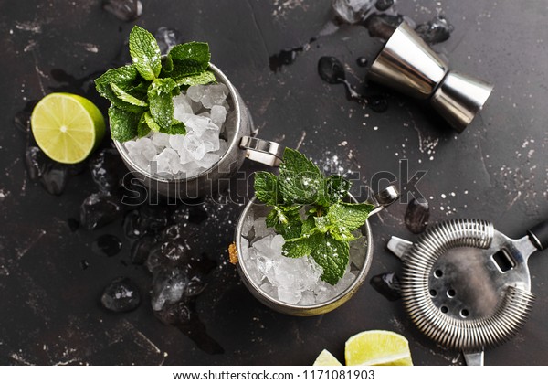 Two\
silver mugs of moscow mule or mint julep cocktail with crused ice,\
mint and lime on dark concrete background. Top\
view