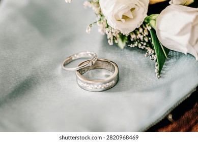Two Silver Wedding Rings Placed Next To White Flowers On A Light Blue Fabric Surface
